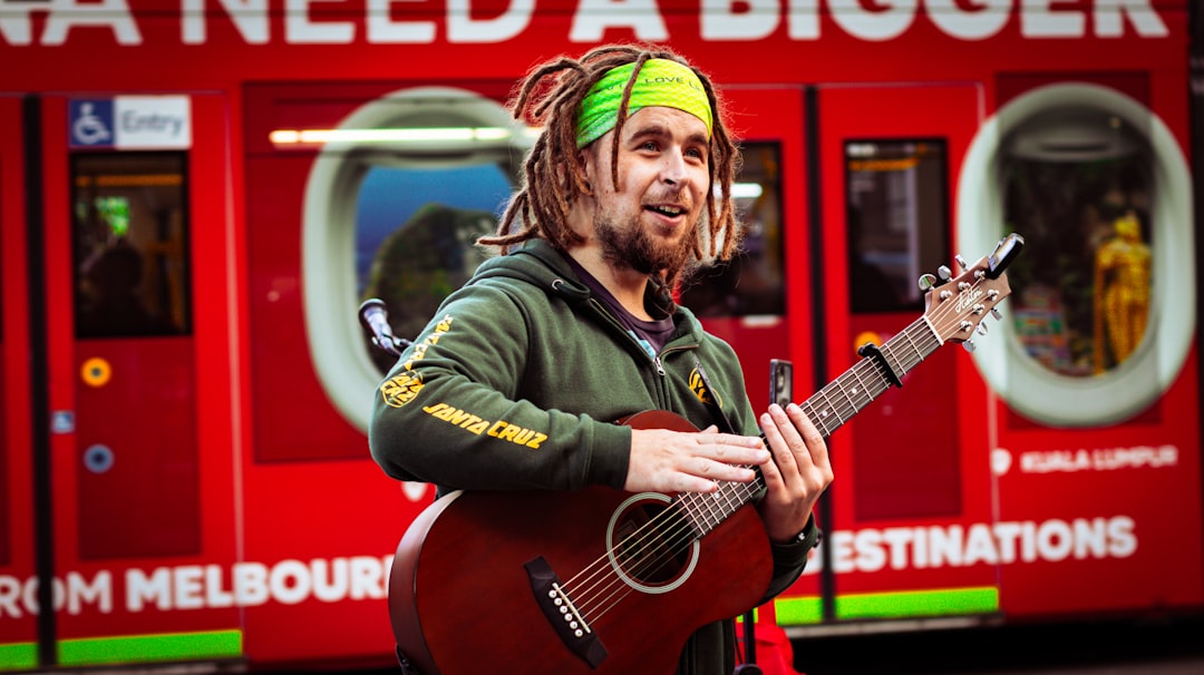 Man with dreadlocks plays acoustic guitar against a red bus.