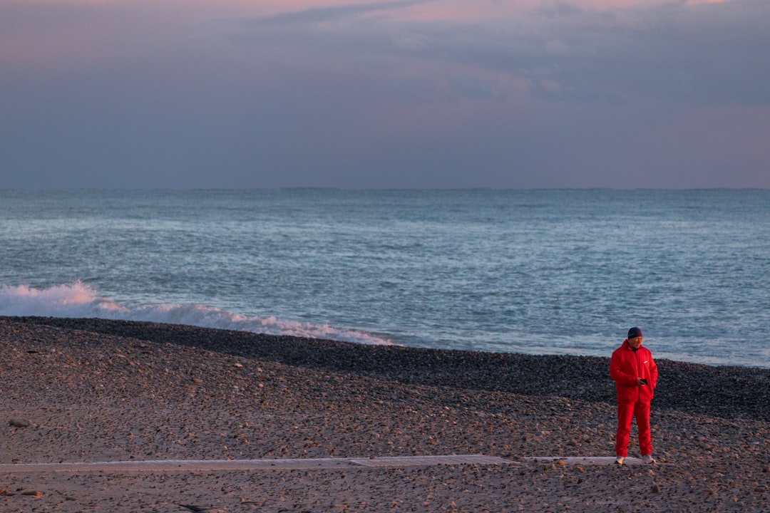 Person in red standing on a pebble beach by the ocean.